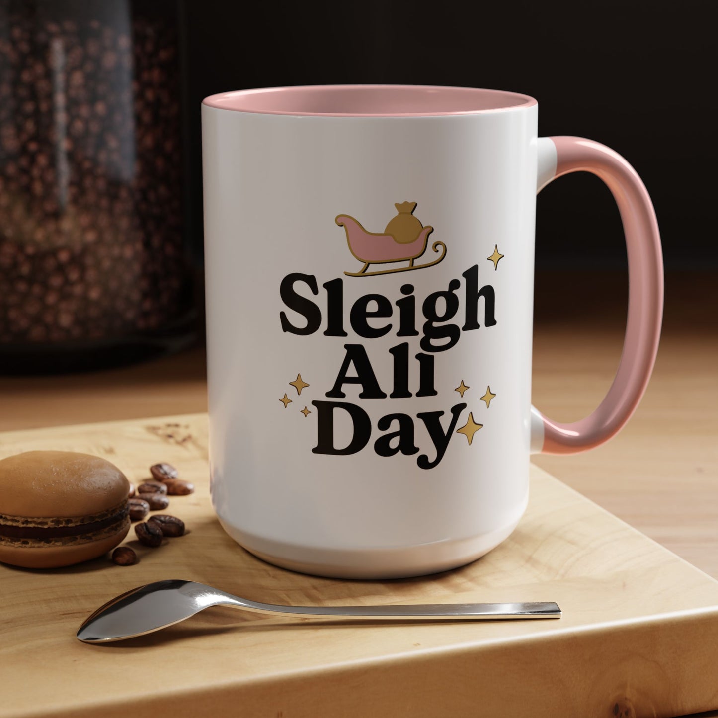 White mug with pink interior and handle, featuring 'Sleigh All Day' text, on a wooden surface with coffee beans and a spoon.