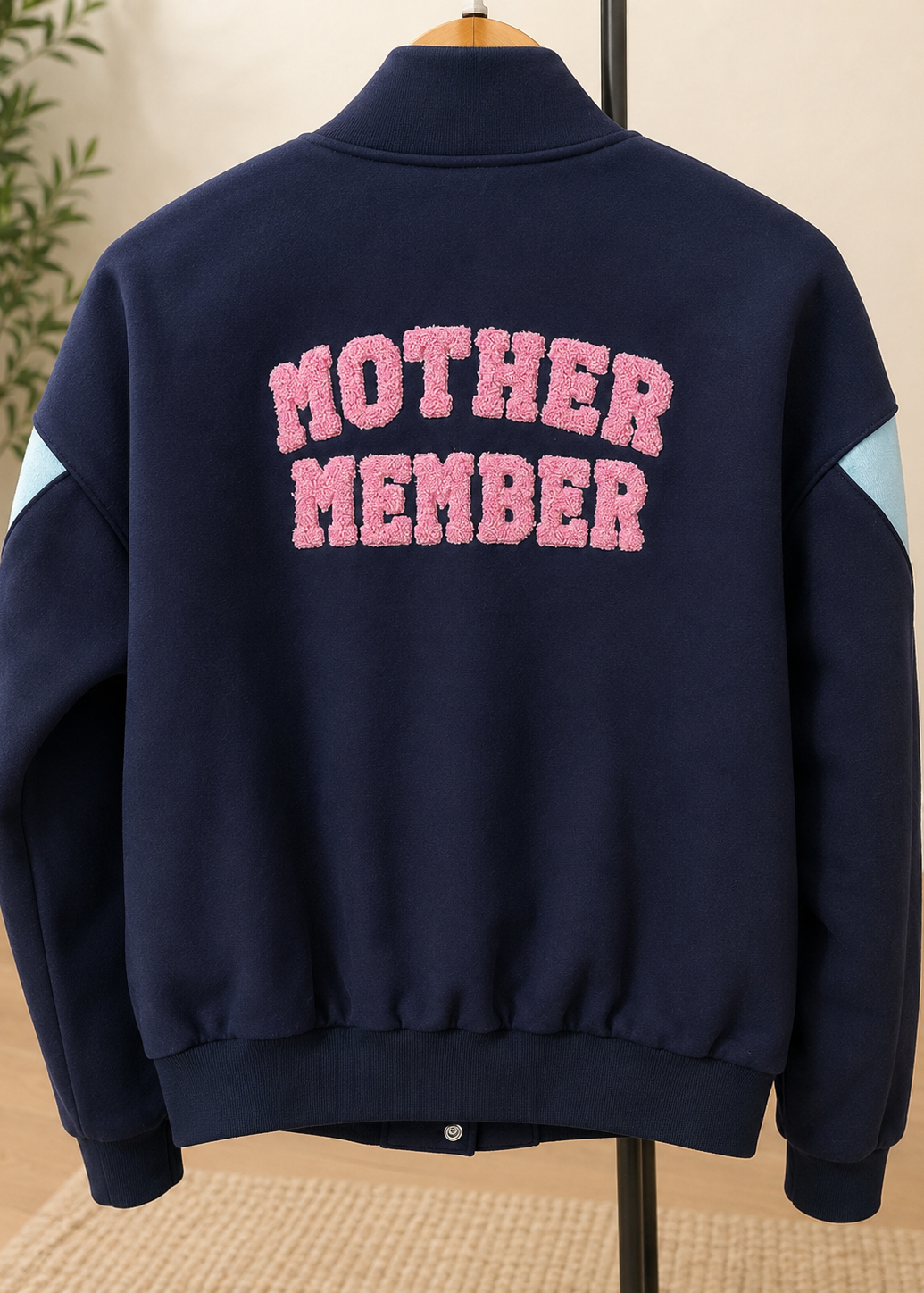 A navy blue fleece jacket displayed on a hanger, featuring the words 'MOTHER MEMBER' in pink fuzzy varsity letters across the back, with light blue sleeve panels.