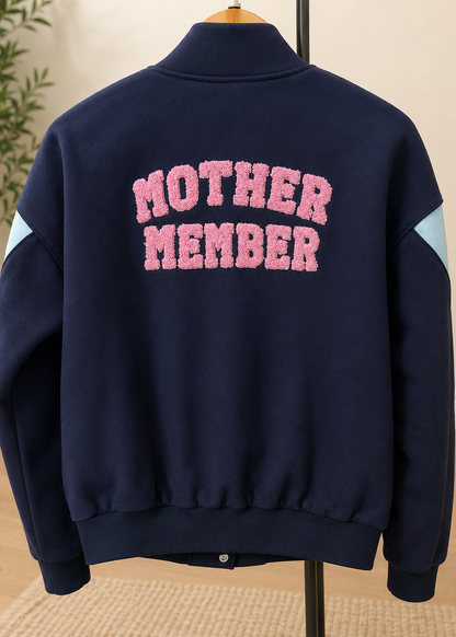 A navy blue fleece jacket displayed on a hanger, featuring the words 'MOTHER MEMBER' in pink fuzzy varsity letters across the back, with light blue sleeve panels.