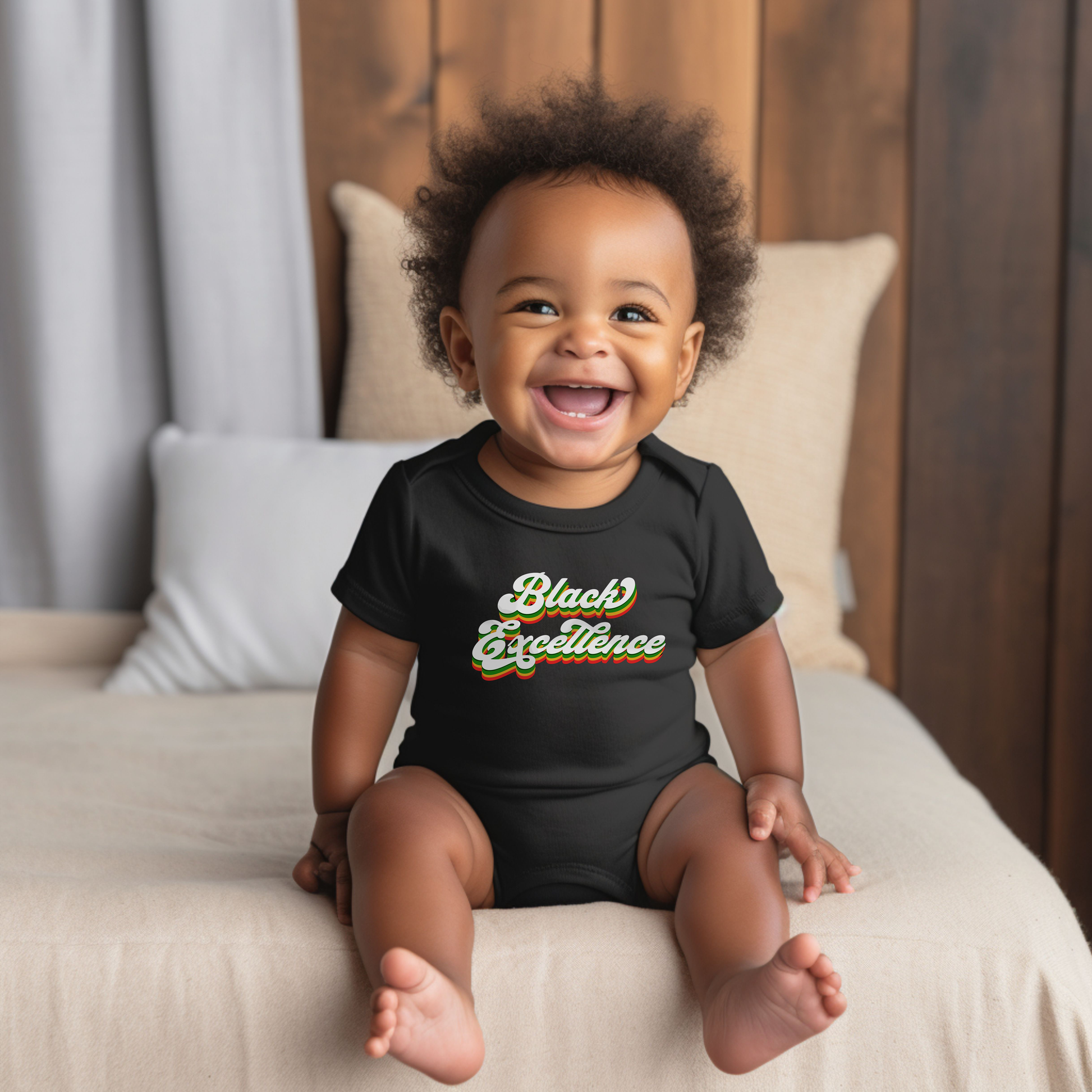 Baby wearing a black onesie with 'Black Excellence' text, sitting on a bed.
