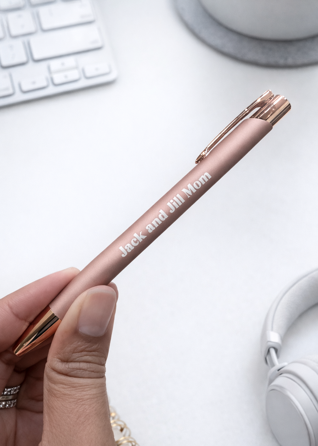 Rose gold pen with 'Jack and Jill Mom' text held by a hand on a white desk with keyboard and headphones.