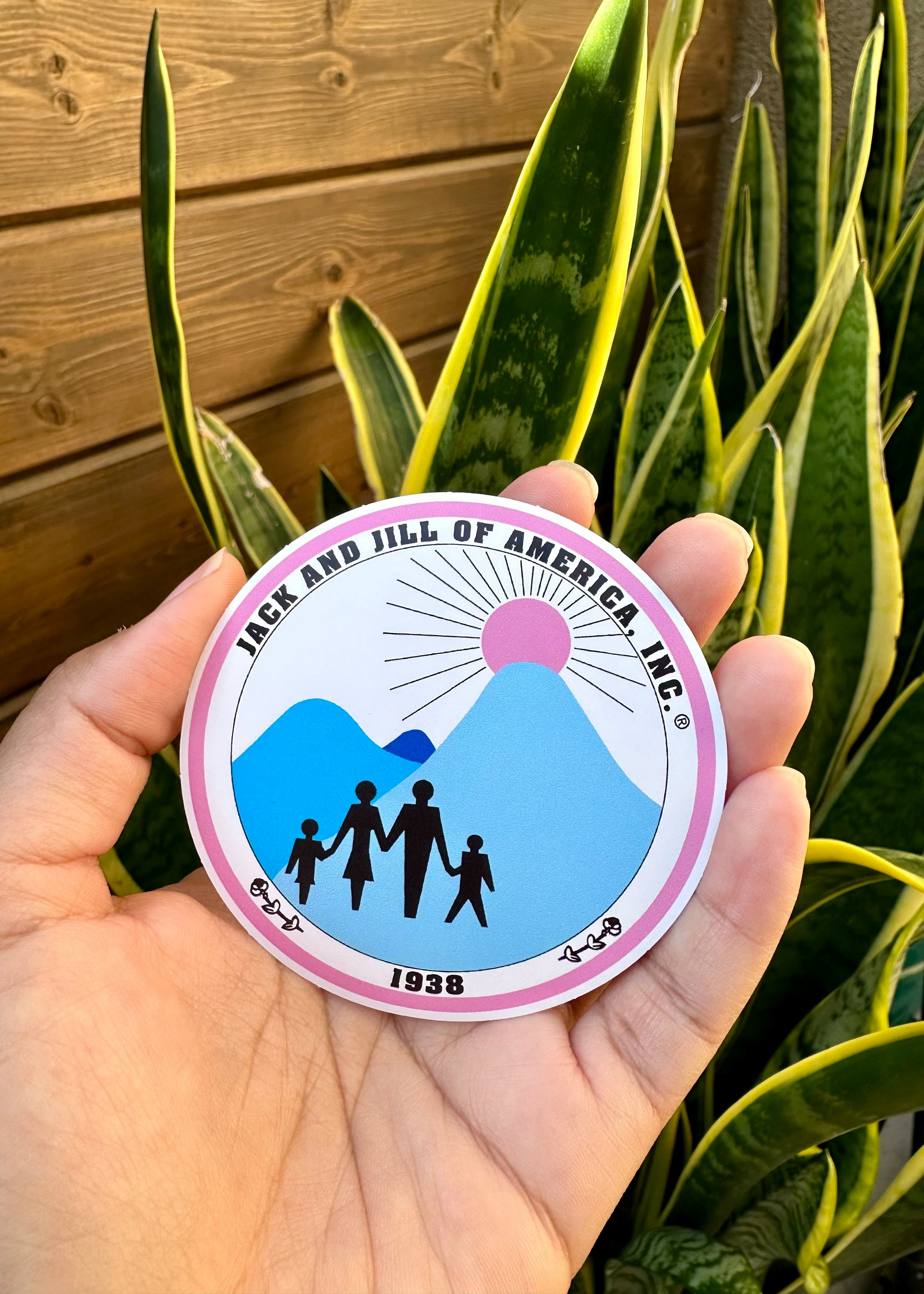 Hand holding a 3 inch circular sticker with 'Jack and Jill of America Inc.' logo against a plant background