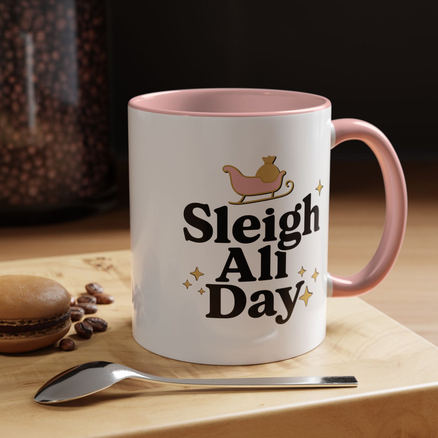 White mug with pink interior and handle, featuring 'Sleigh All Day' text, on a wooden surface with coffee beans and a spoon.