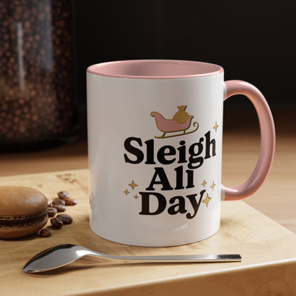 White mug with pink interior and handle, featuring 'Sleigh All Day' text, on a wooden surface with coffee beans and a spoon.